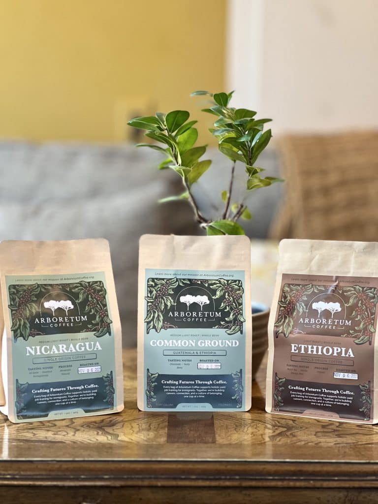 Three bags of coffee labeled "Nicaragua," "Common Ground," and "Ethiopia," placed on a wooden table. In the background, a green plant and a blurred outdoor setting are visible, emphasizing a cozy atmosphere.
