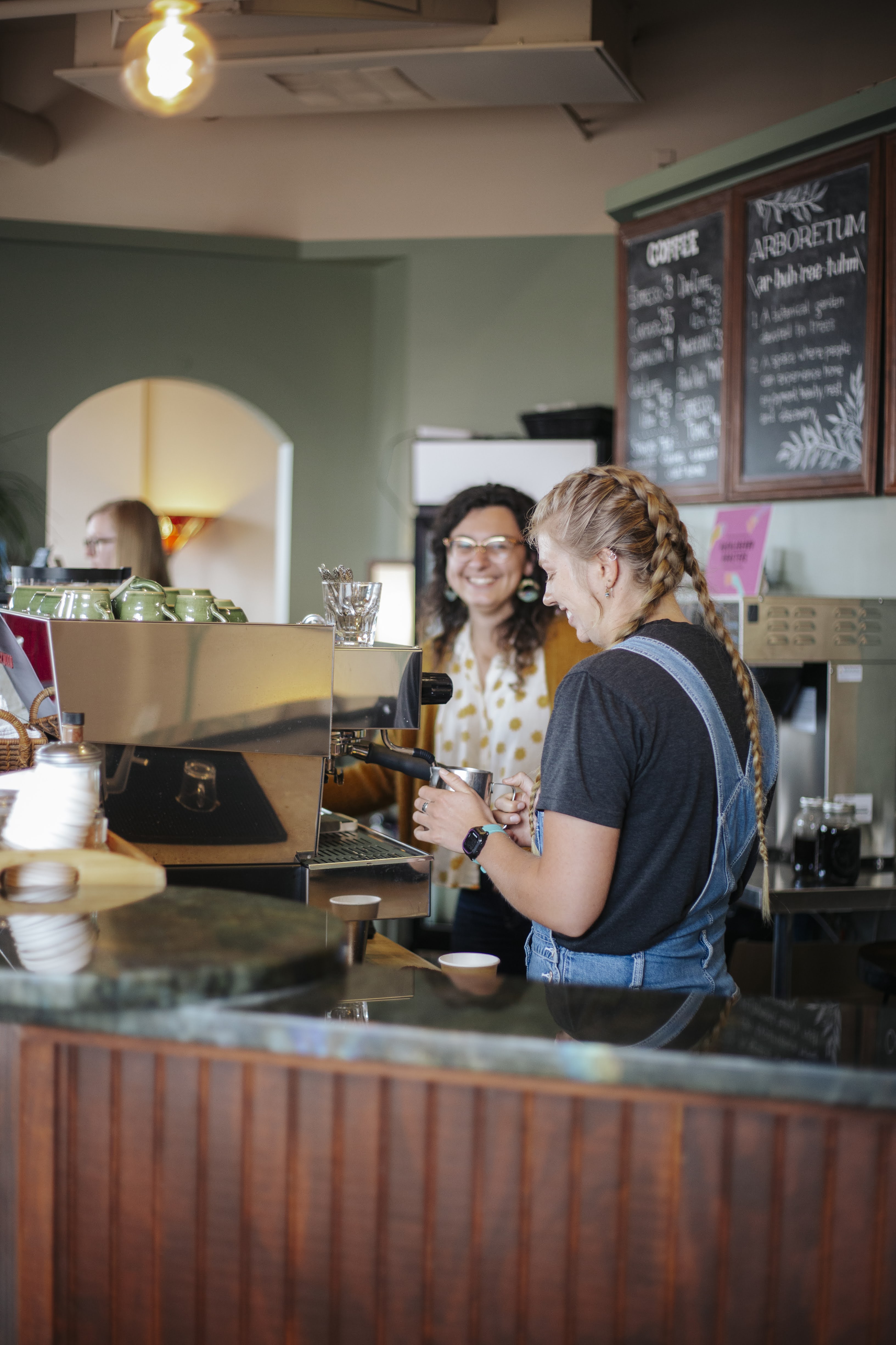 Two women interacting at a coffee shop counter. One woman with glasses smiles while the other, wearing a black shirt and overalls, prepares a drink at the espresso machine. A chalkboard with menu items is visible in the background.