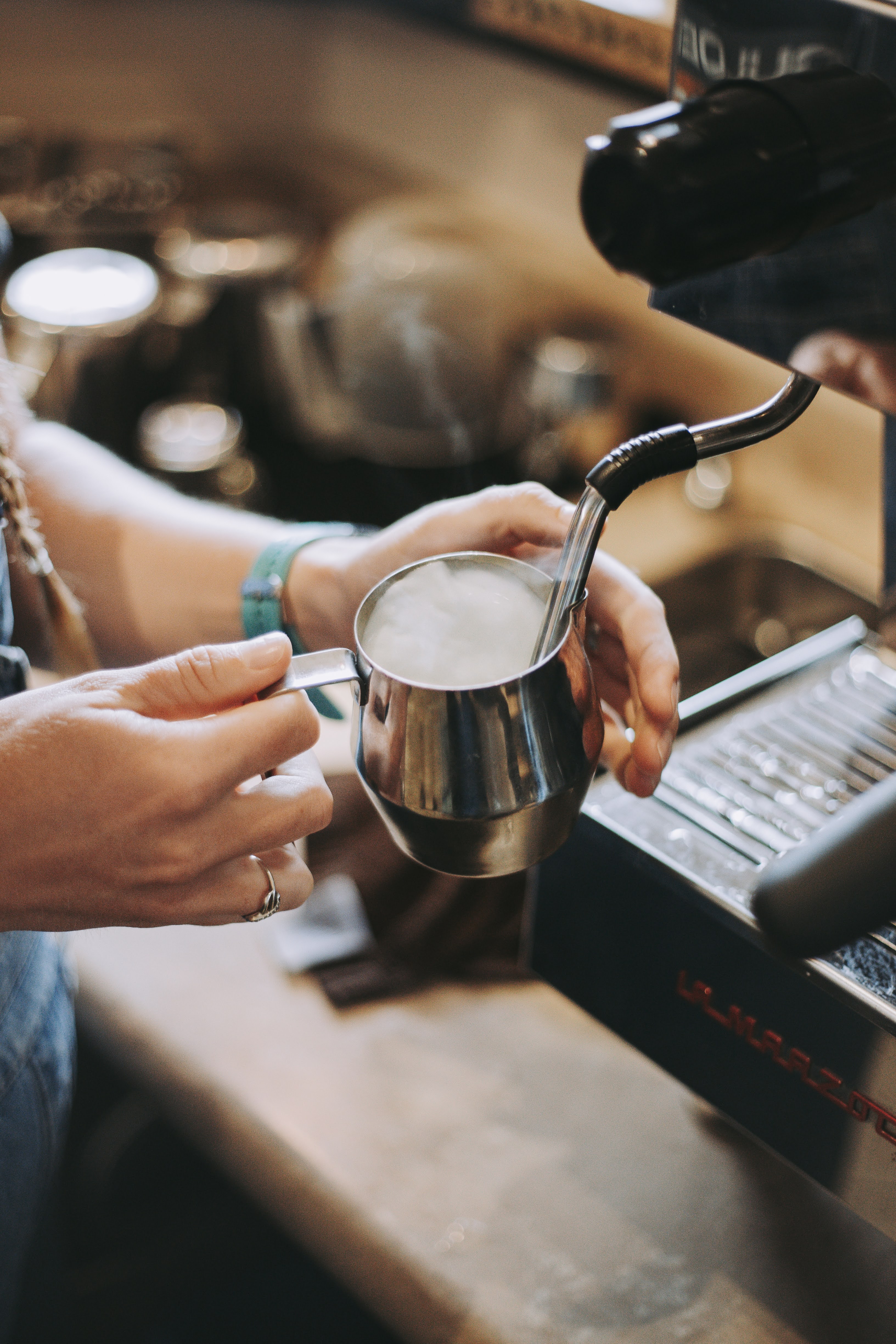 A hand holding a stainless steel milk frothing pitcher near an espresso machine, with blurred kitchen items in the background.
