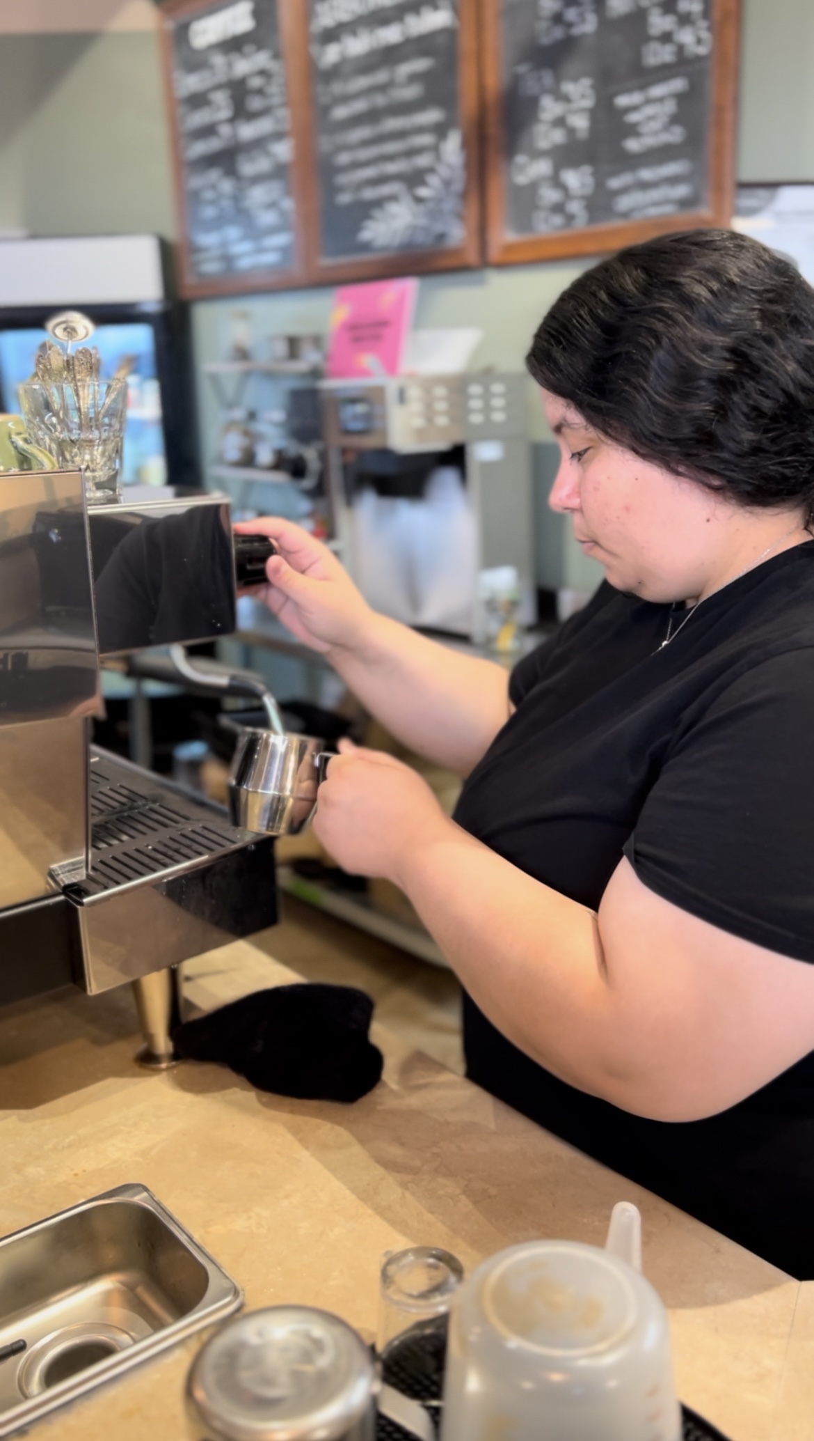 A barista wearing a black shirt is preparing a coffee drink at an espresso machine in a café. The countertop has coffee-making equipment and a focused atmosphere.