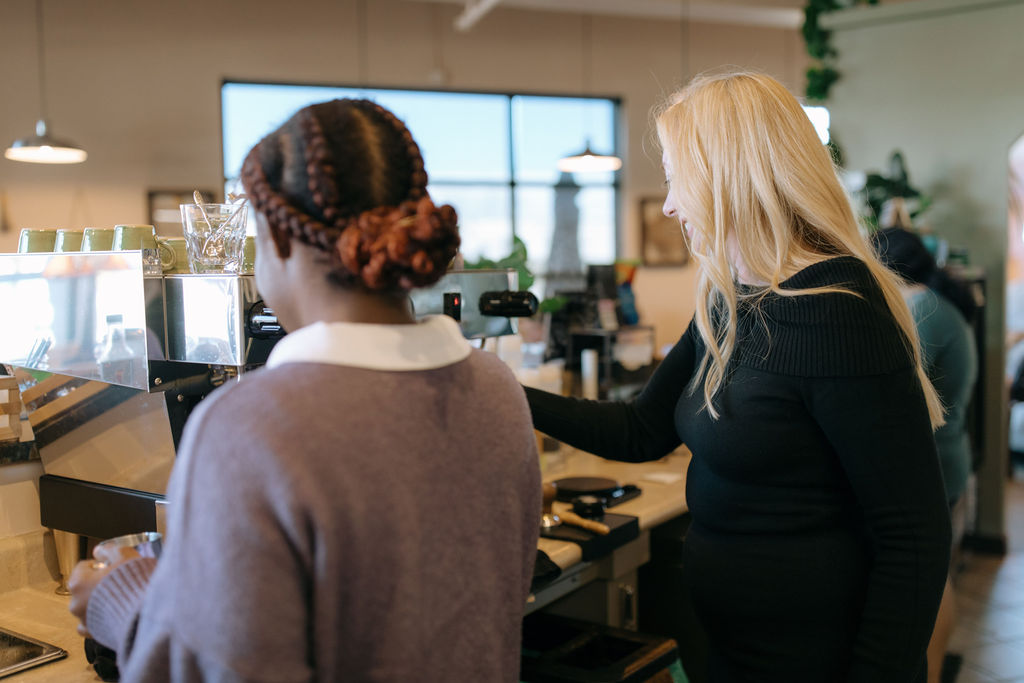 Two women at a coffee shop: one woman with a braided hairstyle operates a coffee machine while the other woman, with long blonde hair, observes, both engaged in conversation. The interior has natural lighting and greenery.