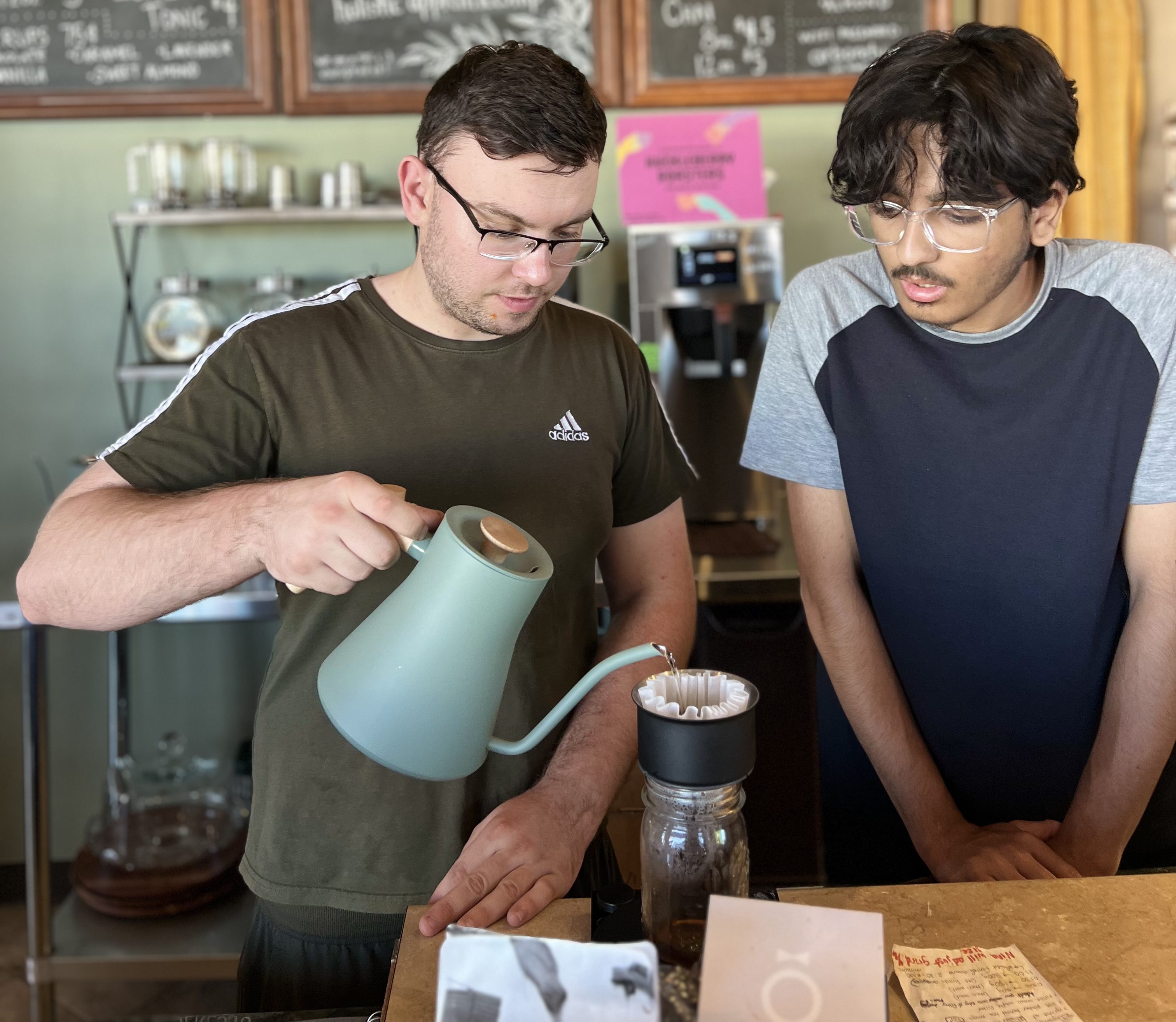A barista in a green shirt pouring coffee from a kettle into a filter, while a customer in a blue and gray shirt watches attentively. A menu board is visible in the background, detailing coffee and non-coffee options.