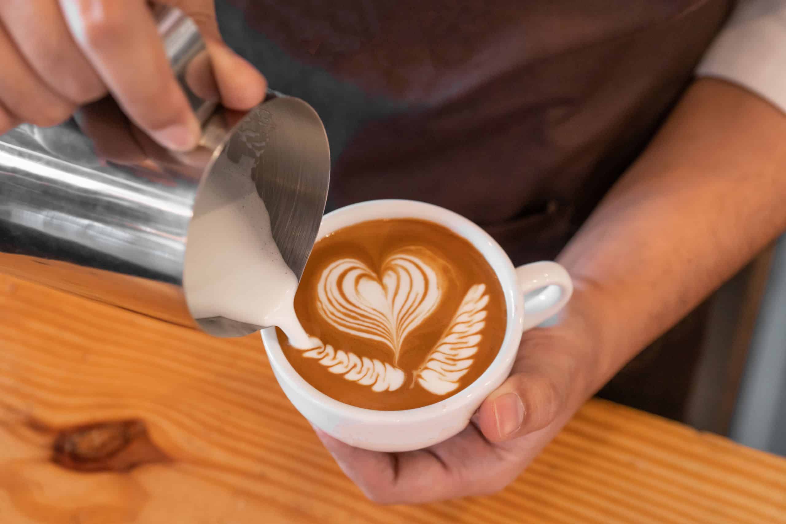 A barista pouring steamed milk into a cup of coffee, creating latte art that resembles a heart and leaves. The cup is placed on a wooden table.