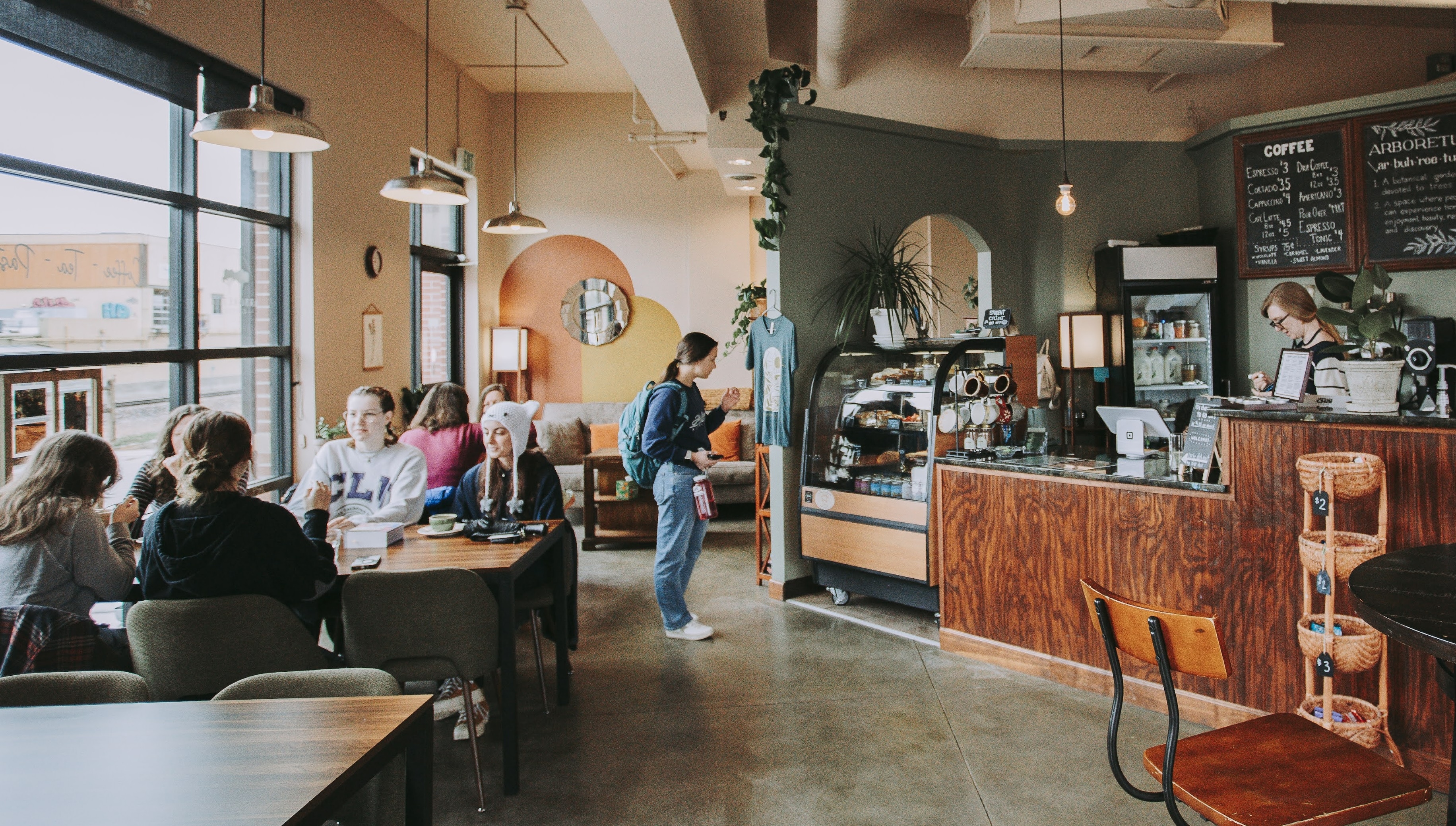 A cozy coffee shop interior with a barista at the counter and several customers seated at tables. Natural light streams through large windows, and plants decorate the space, creating a warm atmosphere.
