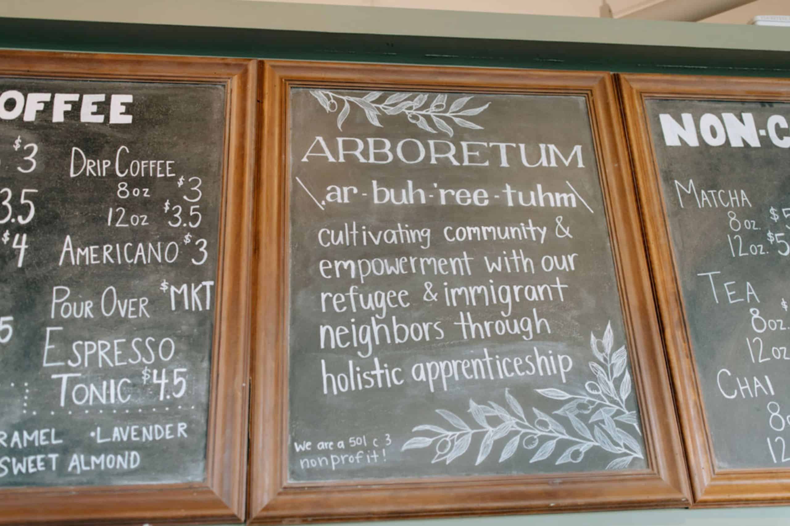 A chalkboard menu displaying coffee and non-coffee options, featuring the word "ARBORETUM" along with its pronunciation and a description about community empowerment and holistic apprenticeship, framed in wood.