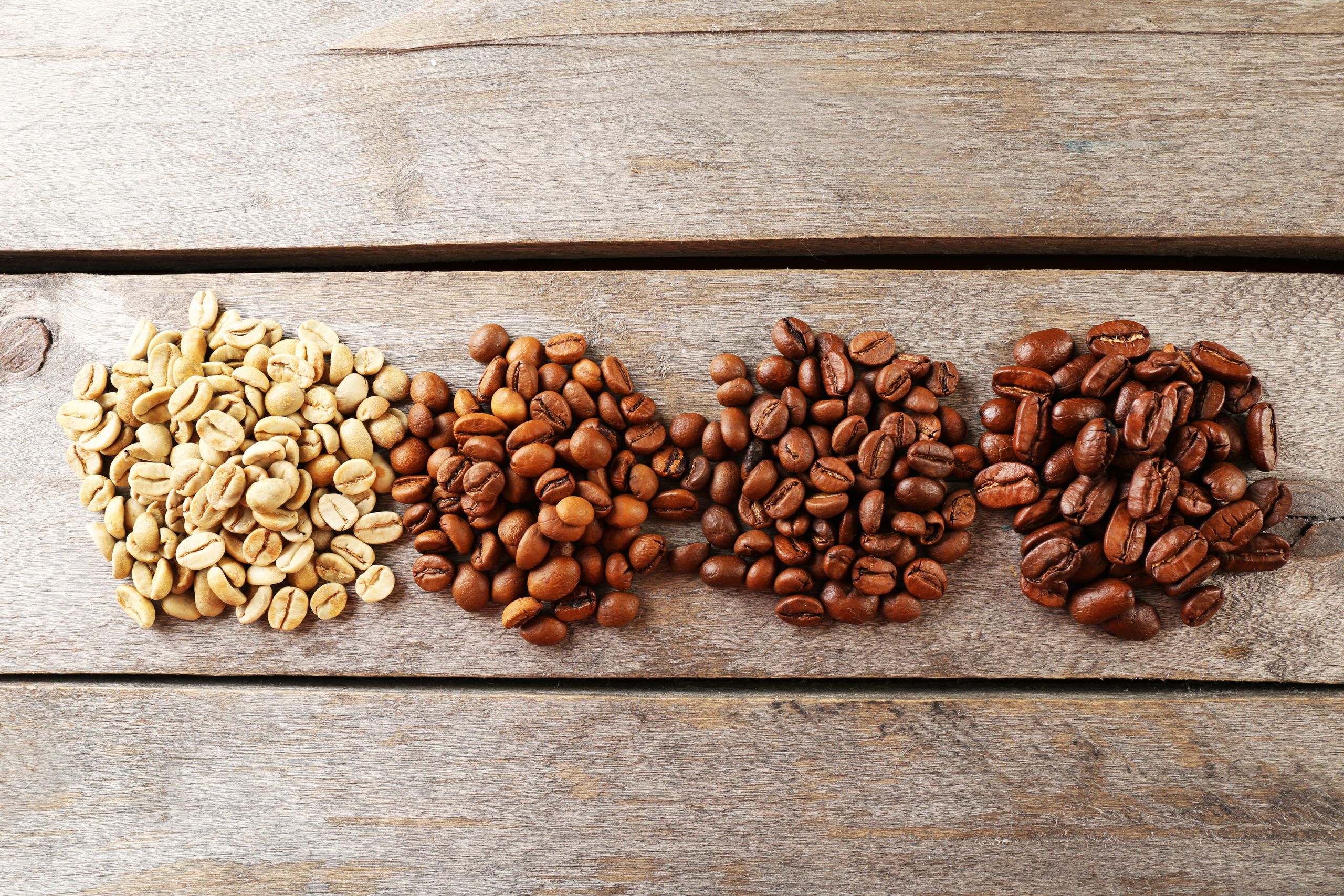 Four types of coffee beans lined up on a wooden surface, showing the stages from unroasted green beans to light, medium, and dark roasted beans.