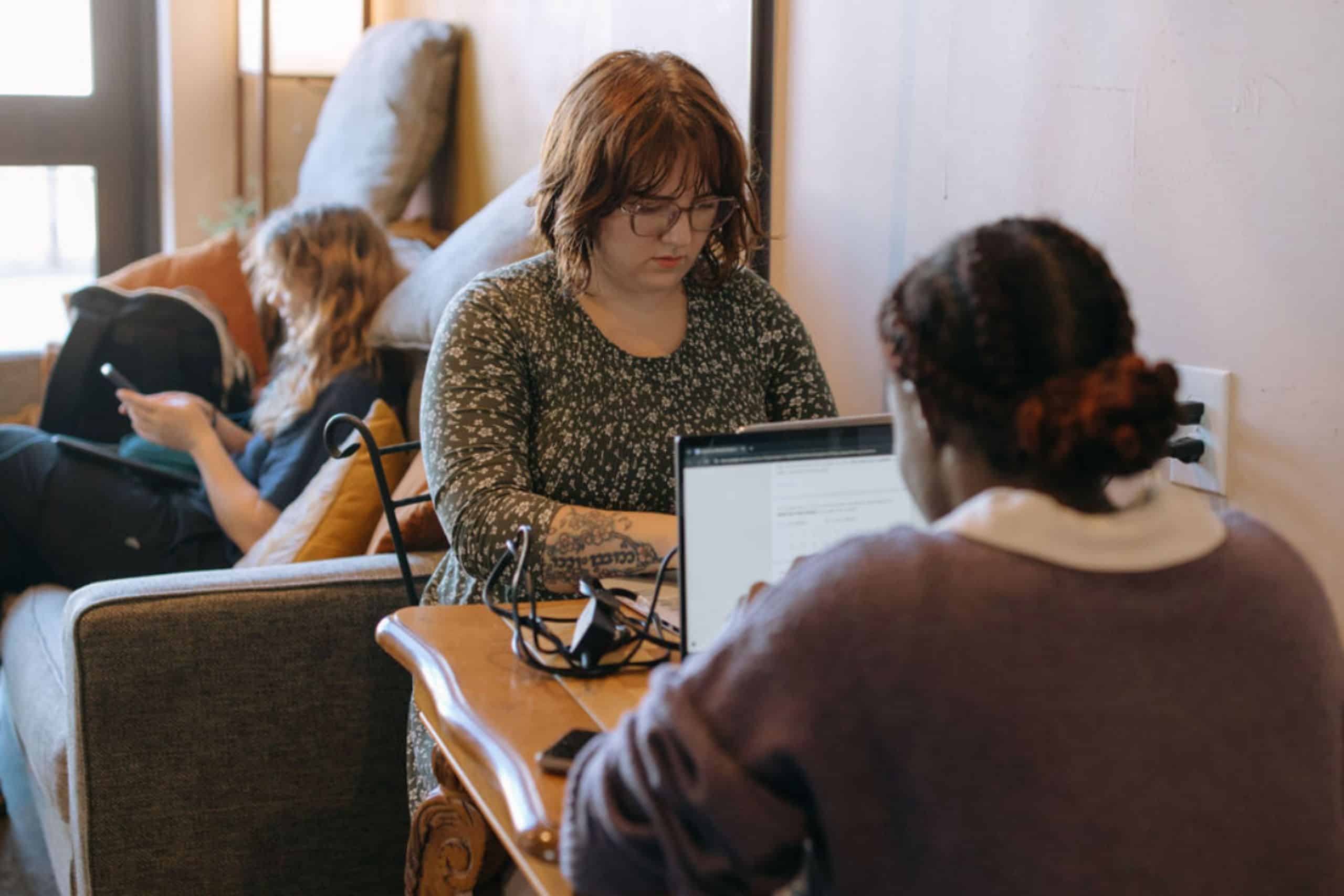 Finding the “Third Place” at Arboretum Coffee 1 Arboretum Coffee Two women working on laptops at a wooden table in a cozy cafe. One woman is focused on her screen, while the other sits on a nearby couch, using her phone. Natural light illuminates the scene.