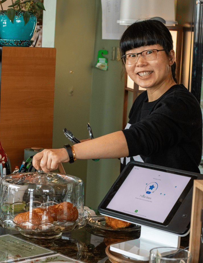 A barista smiling while preparing coffee behind a counter at Arboretum Coffee. Displayed items include pastries under a glass cover, accompanied by a tablet for orders.