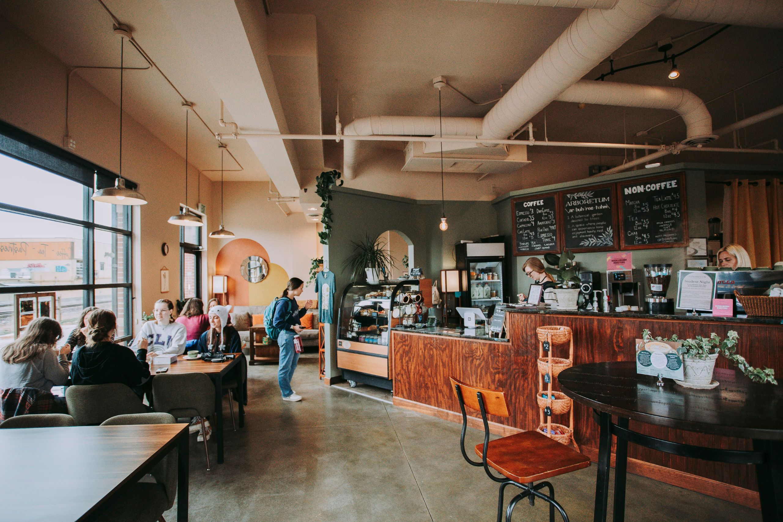 A cozy café interior featuring several tables with customers, a service counter with a barista, and large windows allowing natural light. Decor includes plants and a chalkboard menu on the wall.